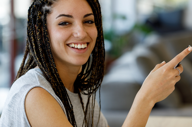 Close-up portrait, blurred background, big teeth, groomed eyebro. Candid close-up portrait of a young woman laughing while using a cell phone, blurred background. She has a nose piercing, big teeth, box braids, groomed eyebrows, possibly freckles, and looks stunning