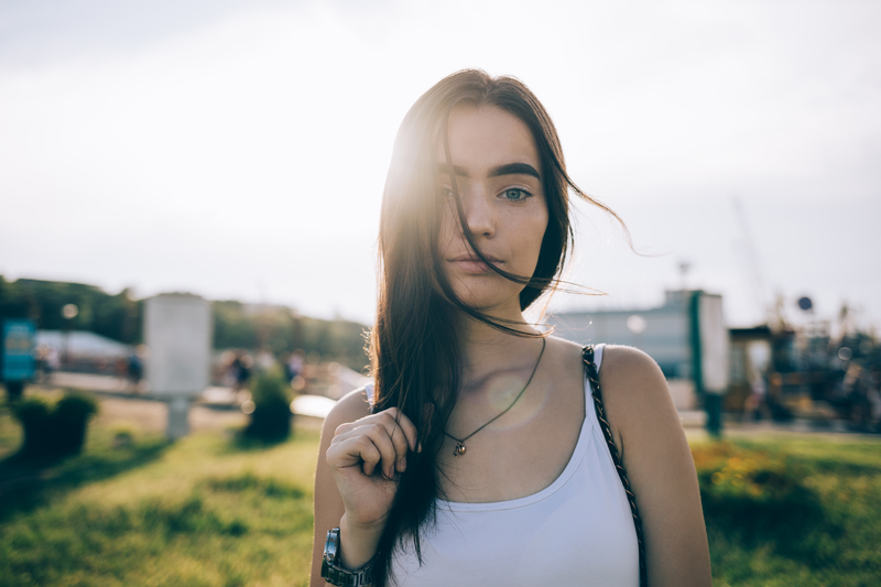 Portrait of stunning young woman wearing white top looking at camera when sun shine from behind. Girl with long brown hair and blue eyes posing in the city park at sunny summer day.